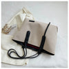 A beige canvas shoulder tote bag with a black strap and a small gold accessory resting on a white surface.
