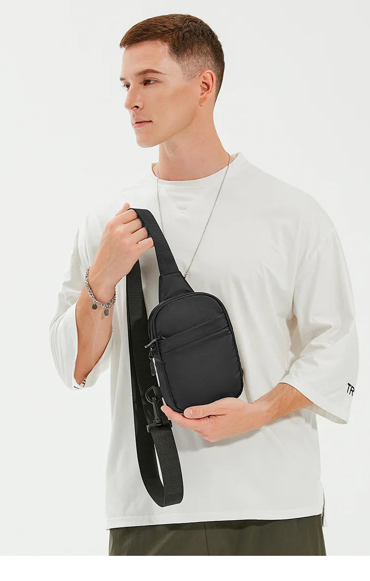 Un modelo masculino sostiene un bolso cruzado negro multifuncional contra una camiseta blanca.