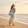 A woman wearing a straw hat stands on a beach with the Turtle Quilted Canvas Tote Bag in her hand.