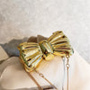 A golden bow knot bag with a detachable gold chain strap resting on a light fabric.
