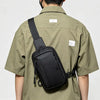 A person wearing a olive green shirt is carrying a black chest bag on their back.