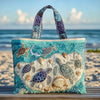 A turquoise quilted canvas tote bag featuring various sea turtle and shell embroidery on a sandy beach.
