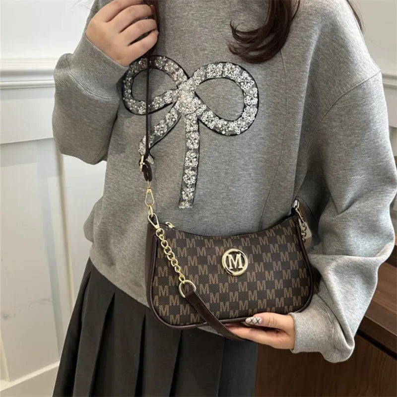 Una mujer con suéter y falda gris sostiene un bolso cruz de cuero negro con cadena dorada y una letra plateada grande
