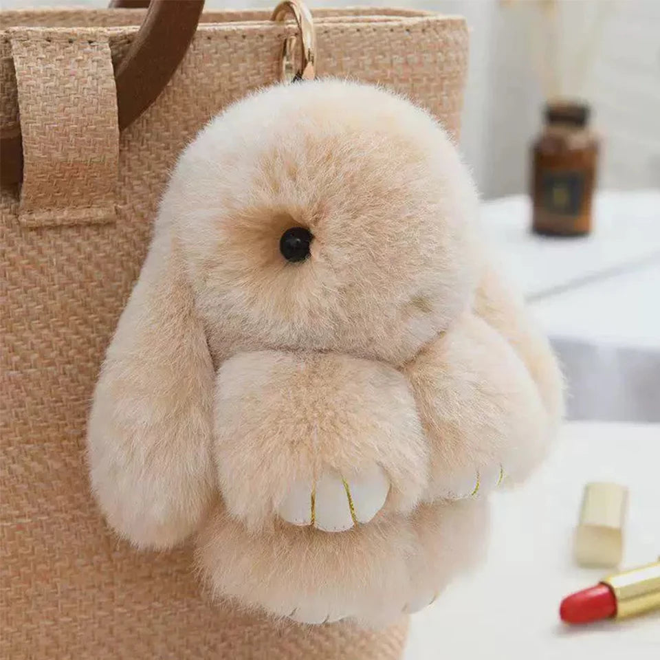 A fluffy, light beige rabbit fur plush keychain resting on a tan canvas bag.
