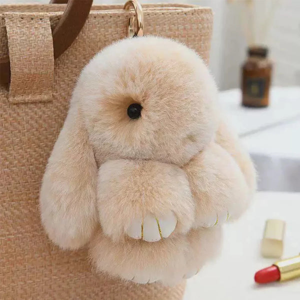 A fluffy, light beige rabbit fur plush keychain resting on a tan canvas bag.