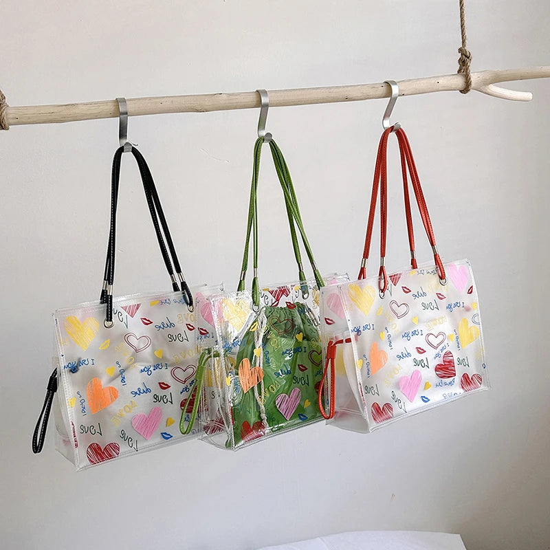 A set of three clear plastic PVC shoulder bags with colorful prints are hanging on a wooden rod.