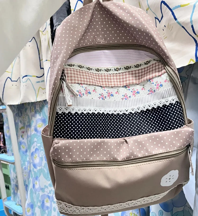 A light pink backpack with floral and polka dot patterns is displayed on a hanger.