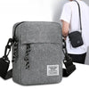 Graue, kompakte Crossbody-Tasche mit schwarzem Riemen und Tasche vor einem weißen Hemd.