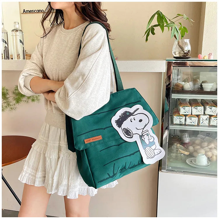 A woman wearing a beige top and white skirt holds a dark green Snoopy canvas tote bag.