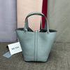 A light blue-green leather bucket bag with a handle is displayed on a gray surface next to other colored bags.