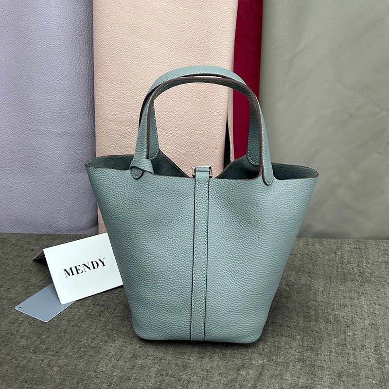 A light blue-green leather bucket bag with a handle is displayed on a gray surface next to other colored bags.