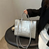 A silver tote bag with leather straps is held by a person against a light background.