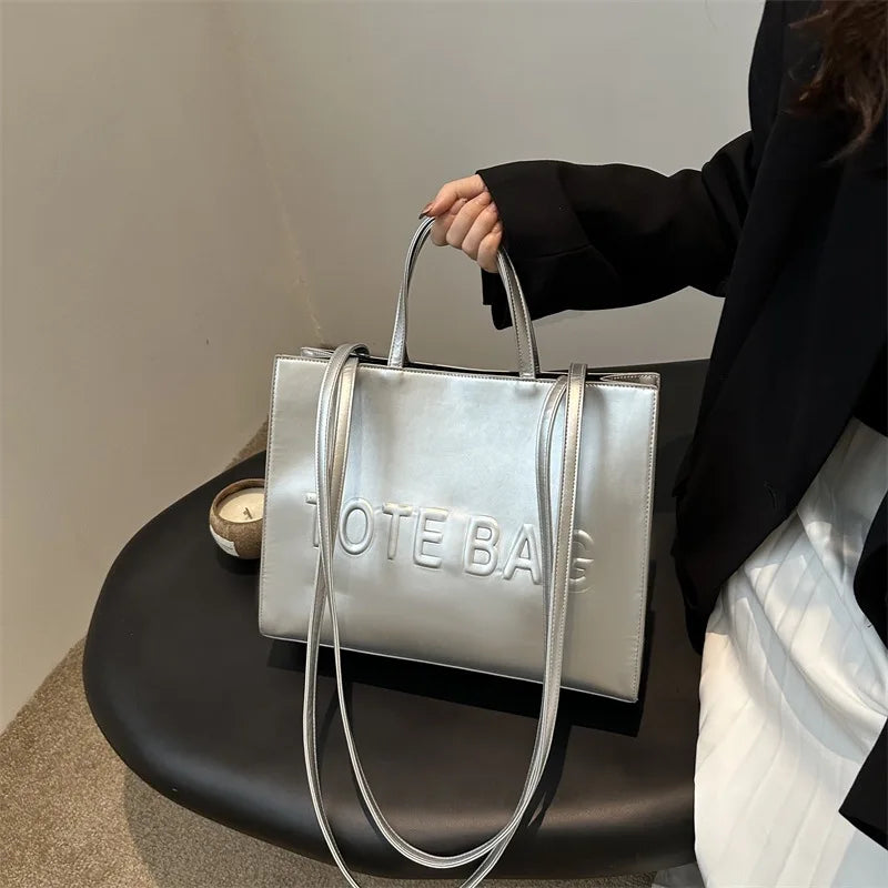 A silver tote bag with leather straps is held by a person against a light background.