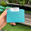 A teal genuine leather zipper card holder with a white tag is held in a hand on a wooden surface.