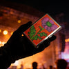 A hand holds an Art Deco metal card holder featuring a vibrant pink and green floral design against a dark background.