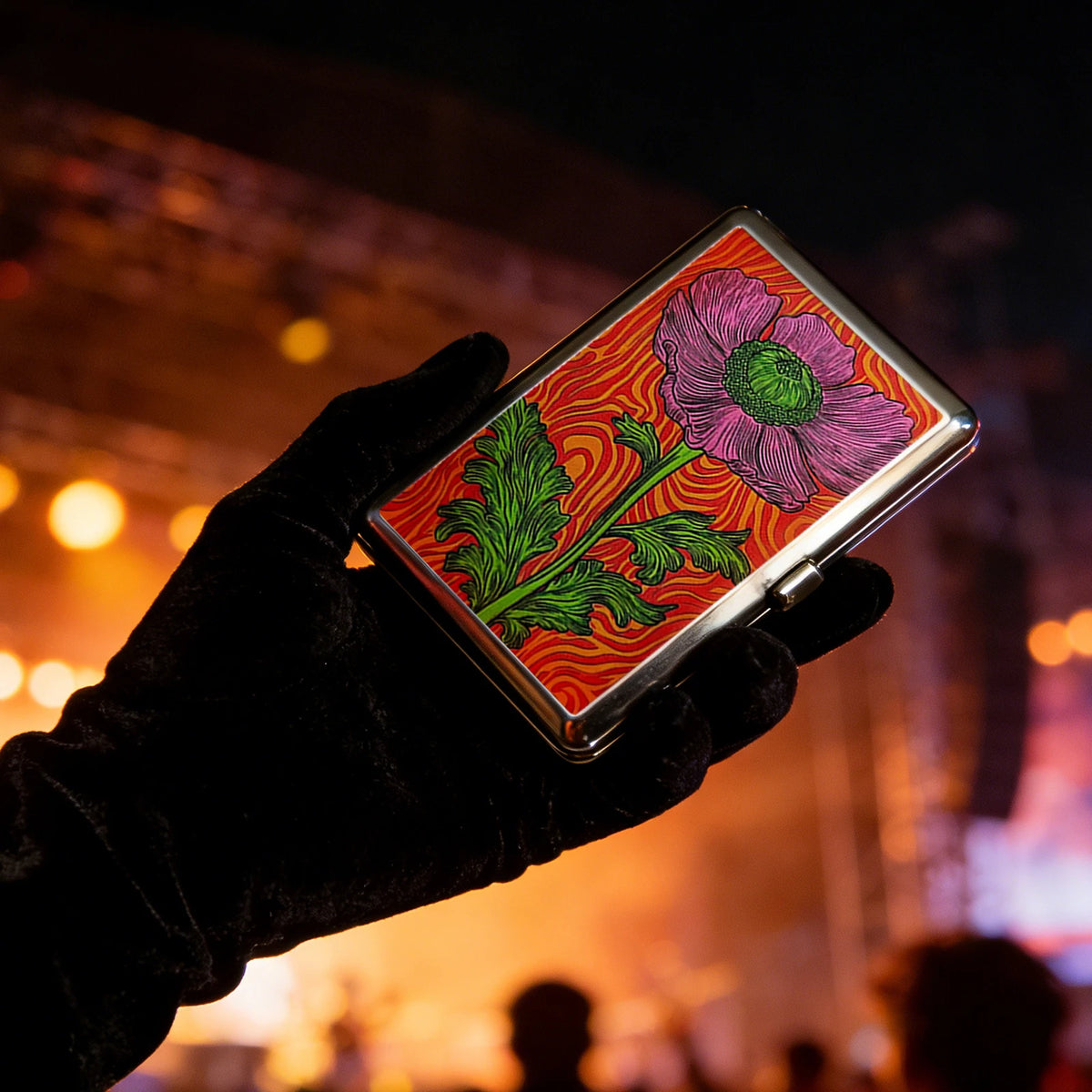 A hand holds an Art Deco metal card holder featuring a vibrant pink and green floral design against a dark background.