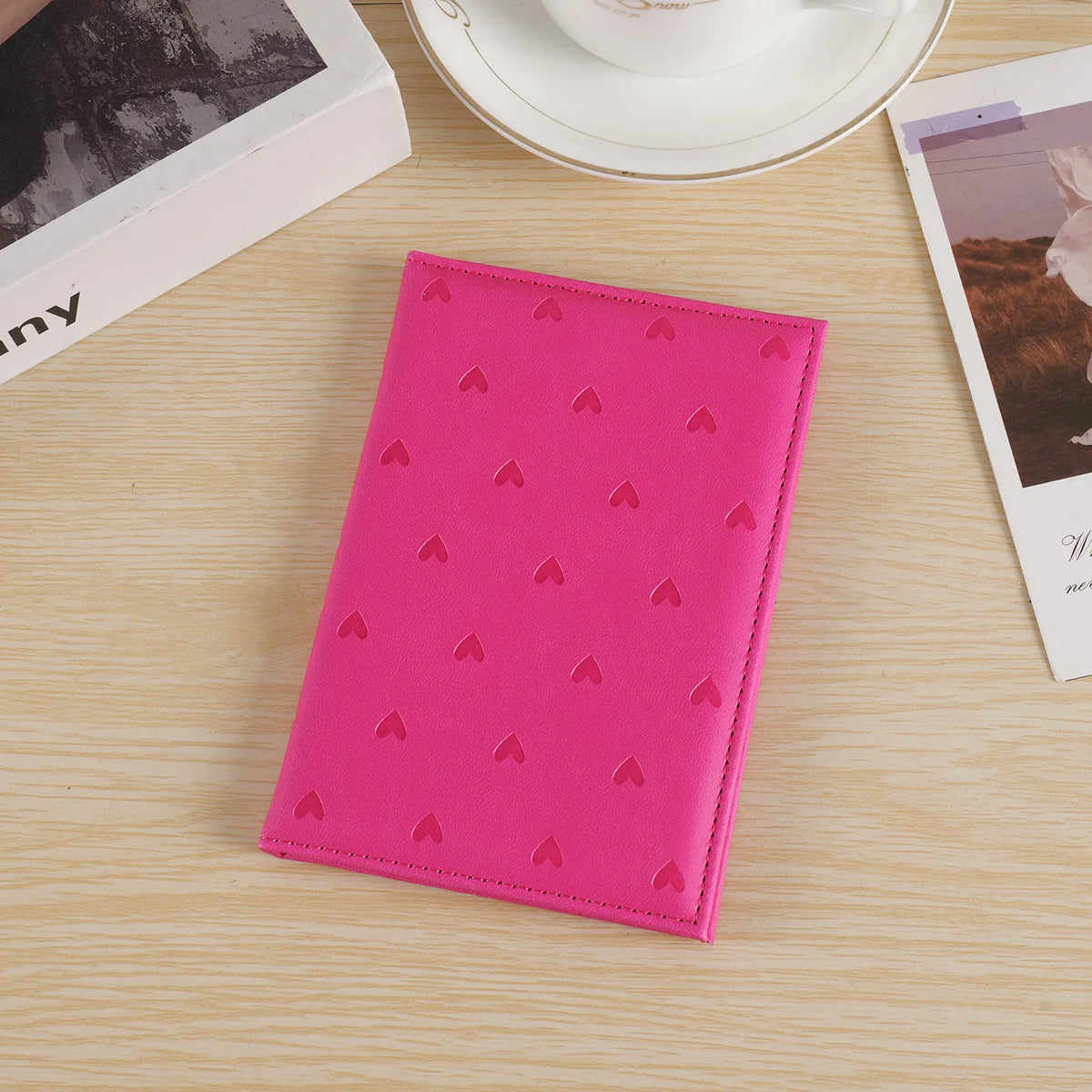 A bright pink heart print passport cover travel wallet resting on a light wooden surface next to a cup and some paper.