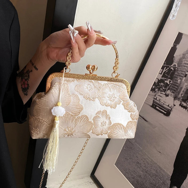 A hand holds an embroidered silk evening clutch with a gold chain strap against a light background.
