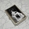 A vintage snap metal card holder displaying a black and white photograph of a draped figure.