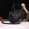 A black crocodile pattern luxury handbag resting on a light surface next to a piece of driftwood.