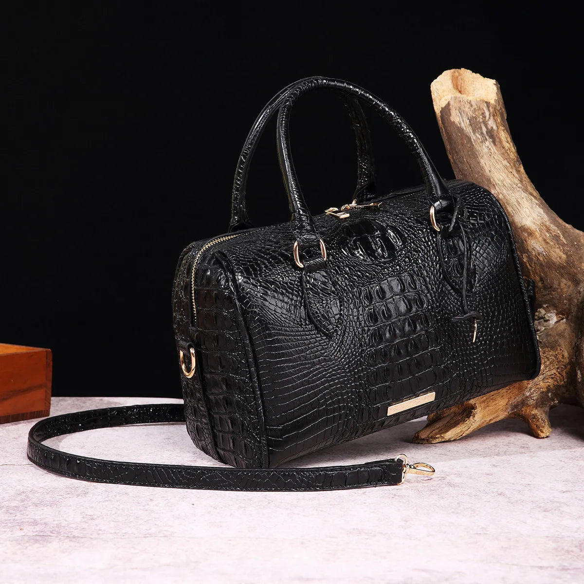 A black crocodile pattern luxury handbag resting on a light surface next to a piece of driftwood.