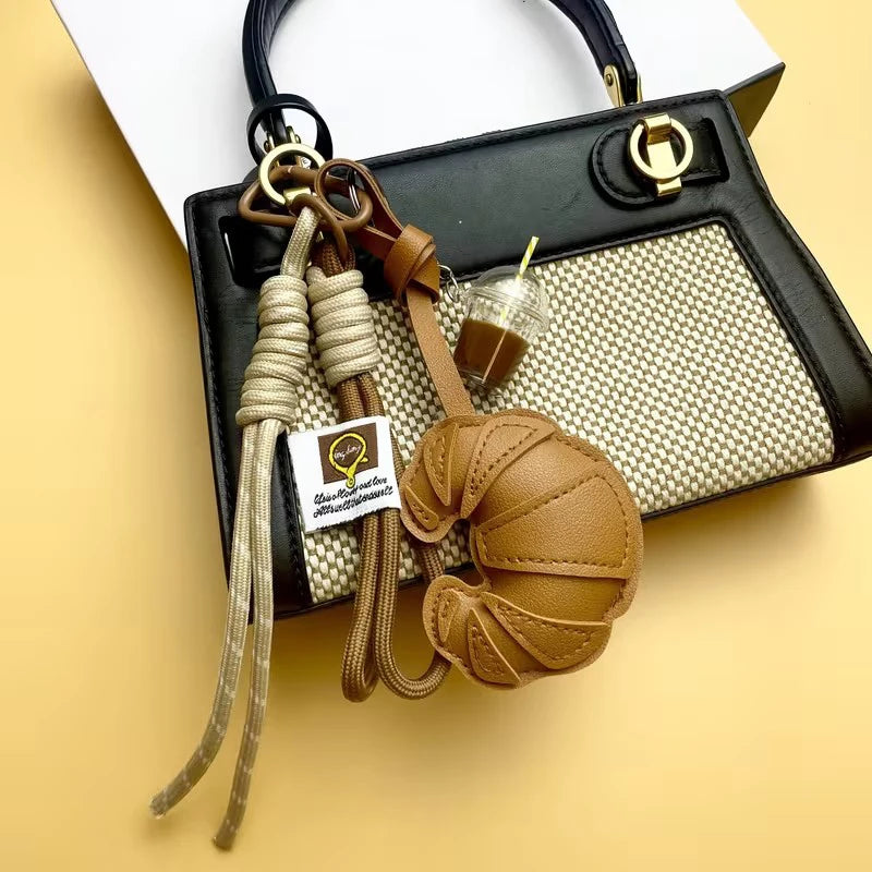 A brown leather croissant keychain with a woven lanyard is displayed against a black and beige handbag.