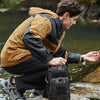 A man wearing tactical outdoor gear sits on rocks near water with a black chest bag.