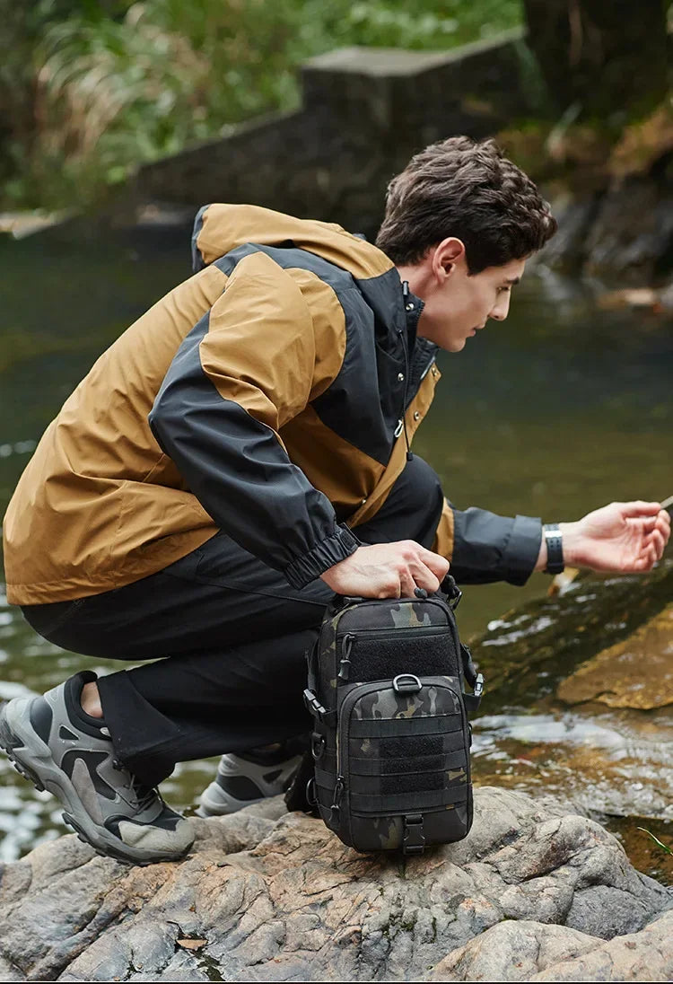 A man wearing tactical outdoor gear sits on rocks near water with a black chest bag.