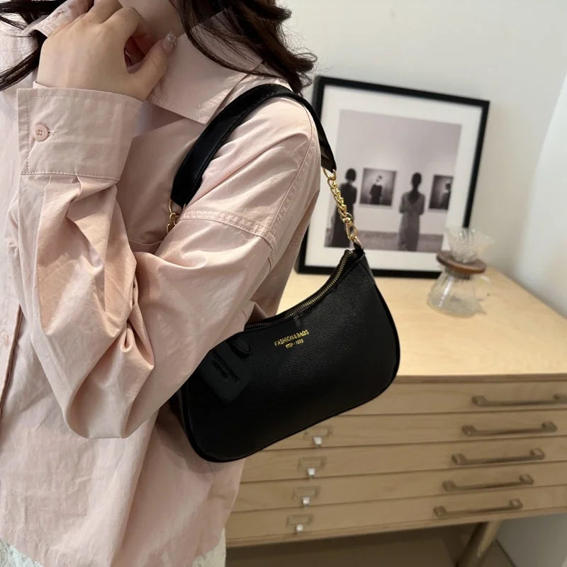 A woman holds a black crescent crossbody bag with a gold chain strap against a light pink shirt.