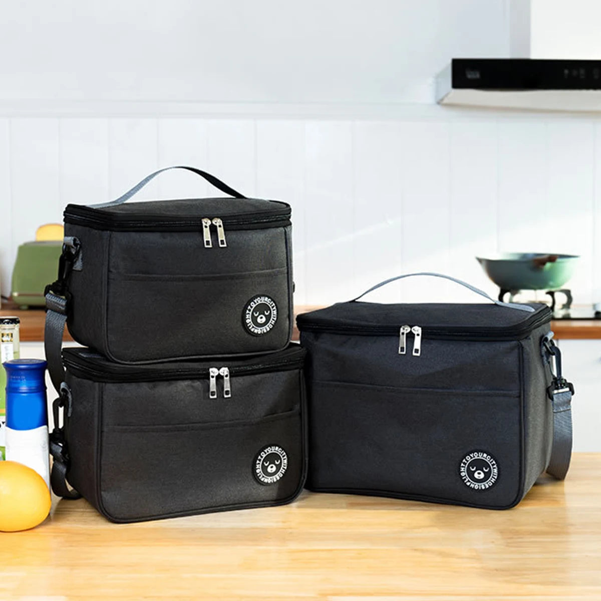A black insulated lunch tote bag with a shoulder strap is displayed on a wooden surface.