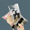 A hand holds a clear plastic pouch containing various makeup brushes and cosmetic tools.