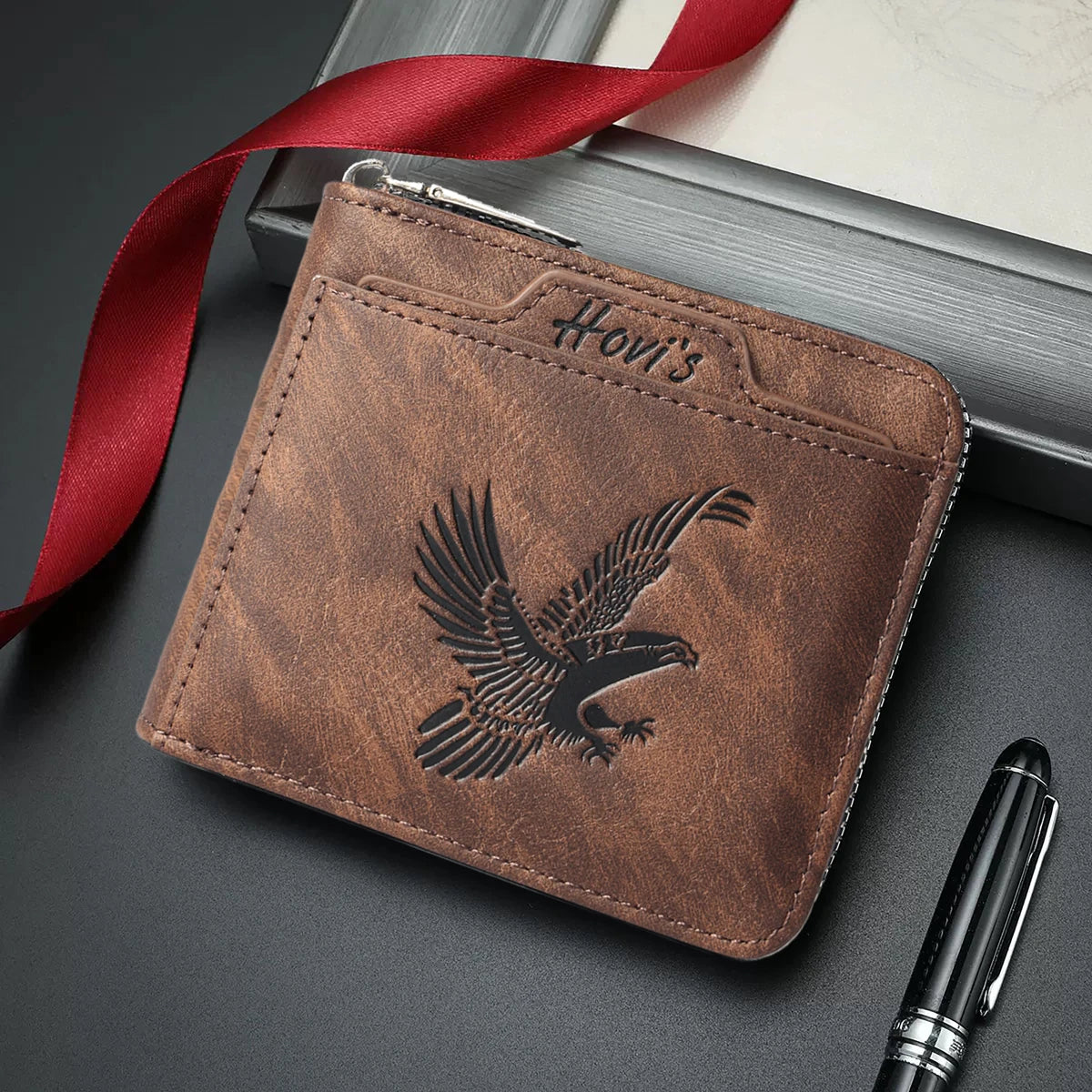 A brown embossed PU leather slim wallet with an eagle design is shown next to a red ribbon and a pen.