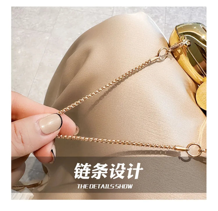 A close-up of a gold chain strap accessory being held against a beige fabric, showcasing the details of the design.