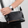 A black everyday crossbody bag with a leather strap and a watch is shown being held by a person.