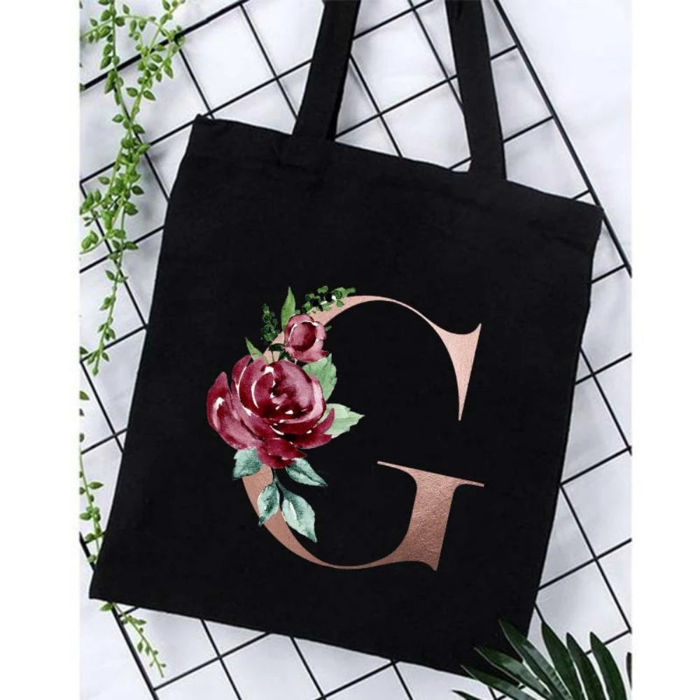 Black canvas tote bag featuring a large gold letter G with floral accents