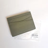 Porte-cartes en cuir véritable vert olive plié dans une gaine en plastique transparent.
