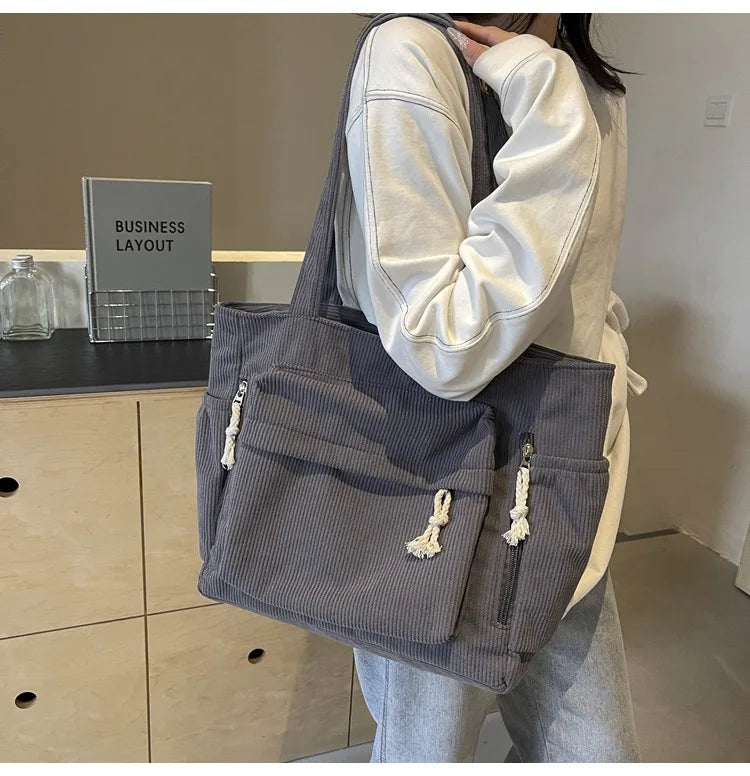 A person holds a gray corduroy shoulder bag with zipper details in a light-colored sweater.