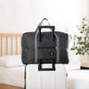 Une valise de voyage noire pliable avec une sangle est posée sur une valise blanche à côté d'un lit.