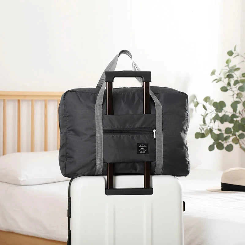 Une valise de voyage noire pliable avec une sangle est posée sur une valise blanche à côté d'un lit.
