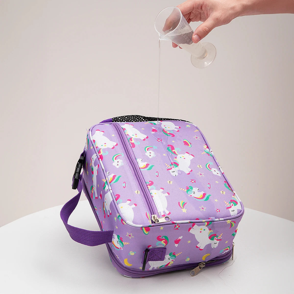 Una bolsa térmica de almuerzo de color morado con patrón blanco y verde se está llenando de líquido de un vaso.