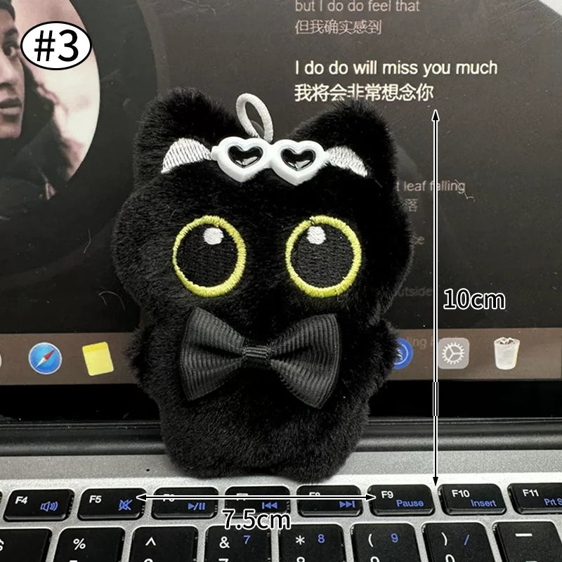 Colgante llavero de peluche de gato negro con ojos grandes y un lazo, apoyado en el teclado de una laptop.