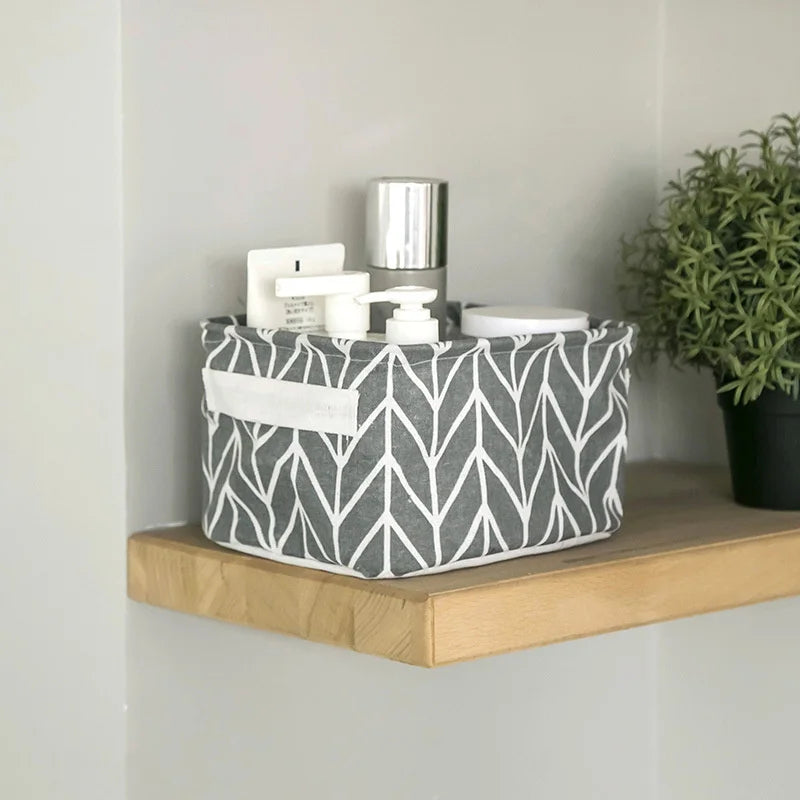 A gray and white patterned foldable cotton linen storage basket with handles holding small white containers on a wooden shelf