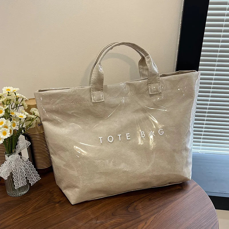 A beige transparent waterproof PVC tote bag is displayed on a wooden surface next to some flowers.