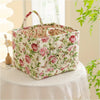 Weiße Blumen-Stoff-Aufbewahrungsbox mit rosa Rosen und grünen Blättern, mit Griffen, auf einer weißen Tischdecke.