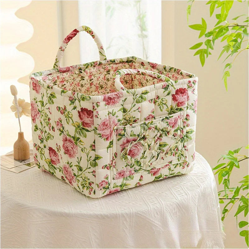 Weiße Blumen-Stoff-Aufbewahrungsbox mit rosa Rosen und grünen Blättern, mit Griffen, auf einer weißen Tischdecke.
