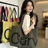 A woman holds a large olive green canvas tote bag with a black logo and shoulder strap in a bright indoor setting.