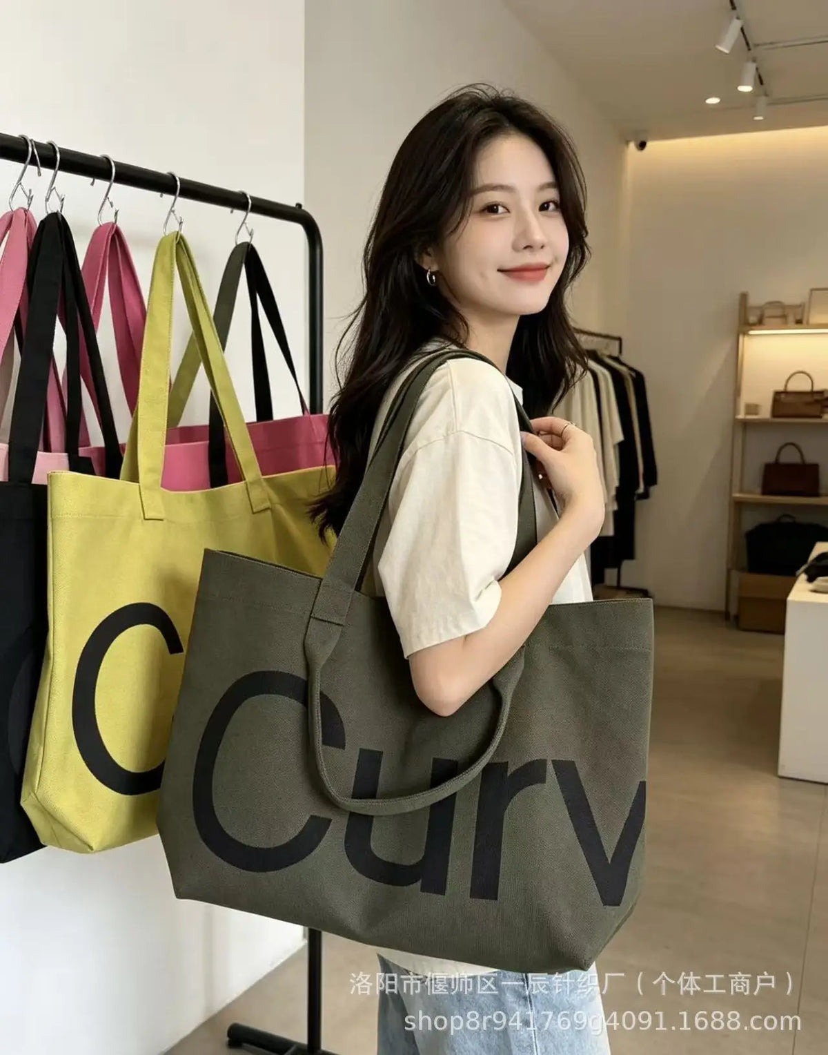 A woman holds a large olive green canvas tote bag with a black logo and shoulder strap in a bright indoor setting.