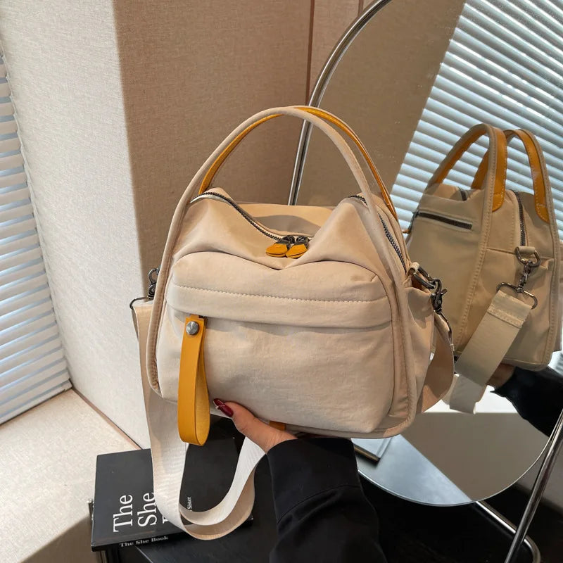 A beige nylon crossbody bag with a top handle is being held by a person against a light background.