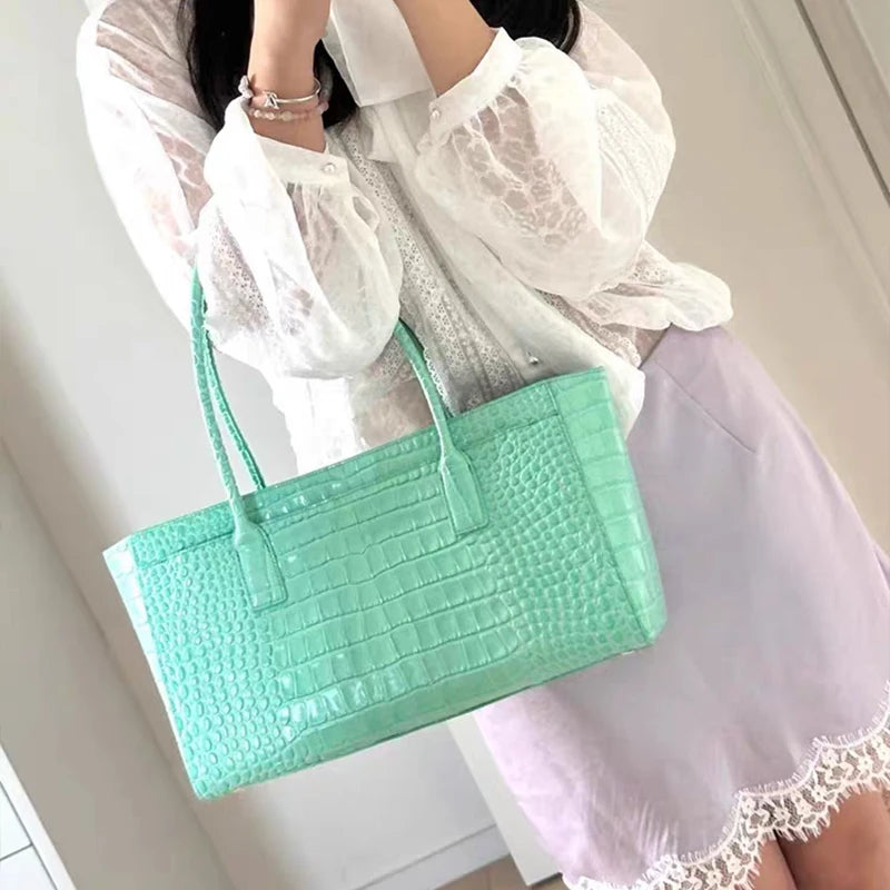 A light mint green crocodile print leather handbag carried by a person wearing a white blouse and lilac skirt.