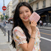 A young woman smiling while holding a pink quilted mini coin purse and card holder wallet outdoors